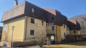 Gelbes Mehrfamilienhaus mit Mansarddach und gepflegter Außenanlage unter blauem Himmel.