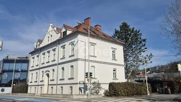 Weißes Altbaugebäude mit rotem Ziegeldach an einer Straßenecke unter blauem Himmel.
