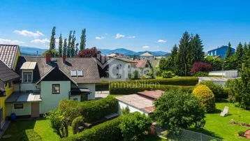 Luftaufnahme von Wohnhäusern mit gepflegten Gärten und Bergblick unter blauem Himmel.