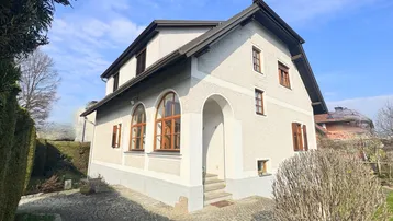 Traditionelles Einfamilienhaus mit heller Fassade, braunen Fensterläden und gepflastertem Eingangsbereich unter blauem Himmel.
