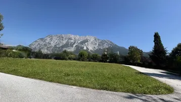 Weitläufige grüne Wiese mit asphaltierter Zufahrt und beeindruckendem Bergblick unter blauem Himmel.