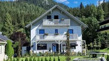 Weißes Haus mit Balkonen und gepflegtem Garten, umgeben von grünen Bergen unter blauem Himmel.