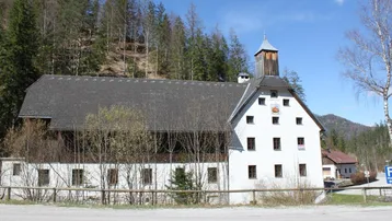 Historisches Gebäude mit weißer Fassade und dunklem Dach, umgeben von Bäumen und Bergen.