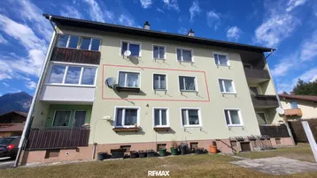 Mehrstöckiges Wohngebäude mit hellgrüner Fassade und Balkonen vor bergiger Landschaft.