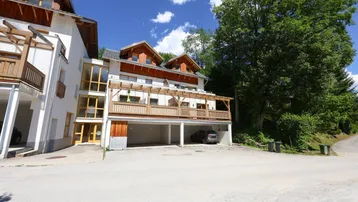 Mehrstöckiges Gebäude mit Holzfassade, Balkonen und überdachten Parkplätzen in grüner Umgebung.