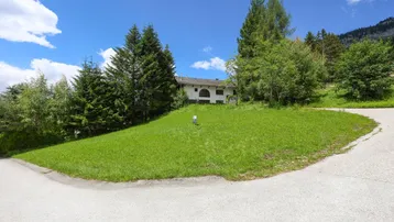 Grünes Grundstück mit sanfter Hanglage, umgeben von Bäumen und einem Haus im Hintergrund, mit Bergblick.