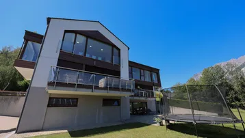 Gartenbereich mit Trampolin und moderner Hausfassade vor Bergkulisse.