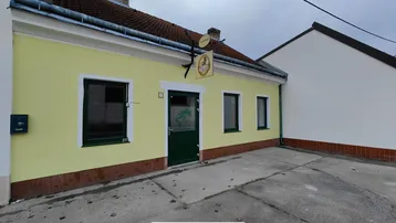 Gelb gestrichenes Gebäude mit grüner Eingangstür und Fenstern, davor ein gepflasterter Vorplatz.