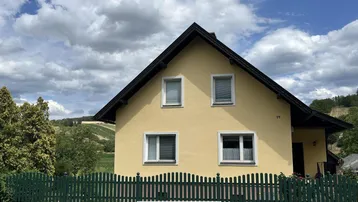 Gelbes Einfamilienhaus mit Satteldach, grünem Zaun und Blick auf die umliegende Landschaft.