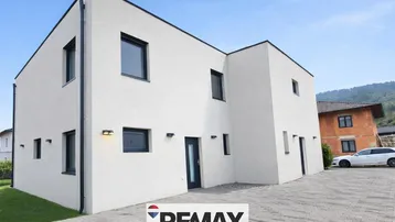Moderne weiße Hausfassade mit klaren Linien und gepflasterter Auffahrt unter blauem Himmel.