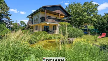 Charmantes Haus mit Balkon und Teich im gepflegten Garten unter blauem Himmel.