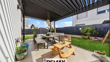 Moderne Terrasse mit Pergola, Gartenmöbeln und kleinem Holztisch, umgeben von Grünflächen.