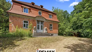 Historisches Gebäude mit Ziegelfassade und Mansarddach, umgeben von Bäumen und trockenem Gras.