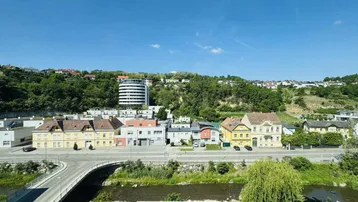 Panoramablick über die Stadt mit Fluss, Brücke und grünen Hügeln unter blauem Himmel.