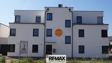 Moderne weiße Fassade eines Mehrfamilienhauses mit dunklen Fenstern und kleinem Balkon.