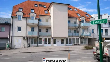 Mehrfamilienhaus mit mehreren Balkonen und Geschäften im Erdgeschoss an einer belebten Straße.