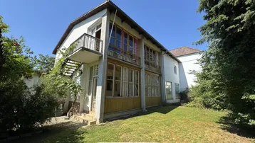 Altes Gebäude mit Balkon und Veranda, umgeben von viel Grün und Bäumen.