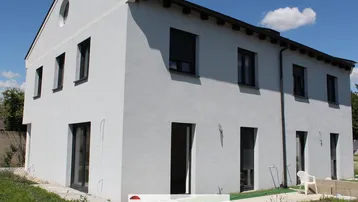 Moderne Hausfassade in Weiß mit dunklen Fensterrahmen und Rollläden, umgeben von Grünflächen.