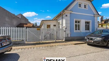 Blaues Einfamilienhaus mit weißem Zaun und gepflastertem Vorgarten an einer ruhigen Straße.