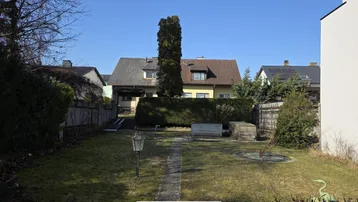 Großer Gartenbereich mit Rasenfläche und Hecken, umgeben von weiteren Wohnhäusern unter blauem Himmel.