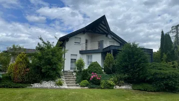 Einfamilienhaus mit gepflegtem Vorgarten, Balkon und markantem Dachgiebel unter bewölktem Himmel.