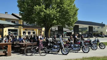 Motorradfahrer und Gäste genießen das sonnige Wetter auf der Terrasse eines Restaurants mit Tankstelle im Hintergrund.