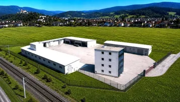 Luftaufnahme eines Gewerbegebiets mit mehreren Hallen und einem Bürogebäude, umgeben von grünen Feldern und Bergen.