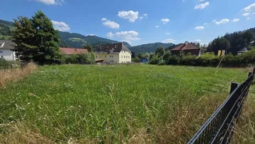 Weitläufiges grünes Feld mit Blick auf umliegende Häuser und Berge unter blauem Himmel.