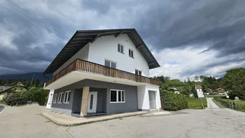 Weißes und graues Haus mit Holzbalkon unter einem dramatischen Himmel an einer Straßenecke.
