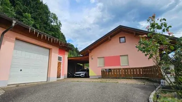 Rosa gestrichenes Haus mit Garage und Carport, umgeben von grüner Natur und Bäumen.