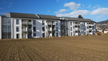Modernes Mehrfamilienhaus mit weißer Fassade, zahlreichen Balkonen und verglasten Treppenhäusern vor ländlicher Kulisse.
