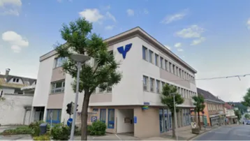 Mehrstöckiges Gebäude mit heller Fassade und blauer Logo an der Ecke, gelegen an einer Straße mit weiteren Häusern.