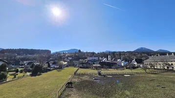 Weitläufiger Blick über grüne Felder, ein Dorf und die umliegende Berglandschaft an einem sonnigen Tag.