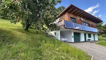 Hausfassade mit Holzverkleidung, Solarpaneelen und integrierter Garage auf einem Hanggrundstück.