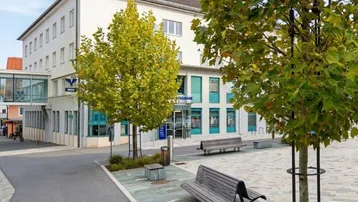 Modernes Gebäude mit öffentlichem Platz, Bänken und Bäumen in einer städtischen Umgebung.