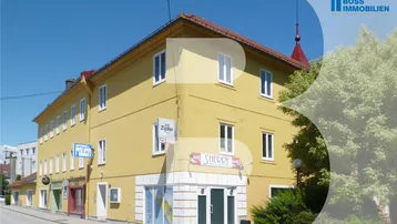 Gelbes Eckgebäude mit Ladenlokalen im Erdgeschoss und Wohnungen darüber an einer städtischen Straße.
