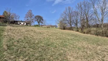 Weitläufiges, leicht abfallendes Wiesengrundstück mit Blick auf entfernte Häuser unter blauem Himmel.