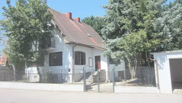 Weißes Einfamilienhaus mit Satteldach, umgeben von Bäumen und einem kleinen Vorgarten.