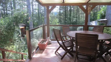Geräumige überdachte Terrasse mit Holztisch und Stühlen, umgeben von üppigem Grün und Bäumen.