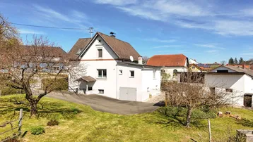 Weißes Einfamilienhaus mit Satteldach und integrierter Garage, umgeben von einem grünen Garten.
