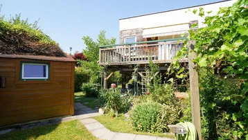 Gepflegter Gartenbereich mit Gartenhaus, Holzterrasse und üppiger Vegetation unter blauem Himmel.