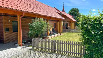 Holzverkleidetes Haus mit rotem Ziegeldach, umzäuntem Garten und gepflastertem Parkplatz.