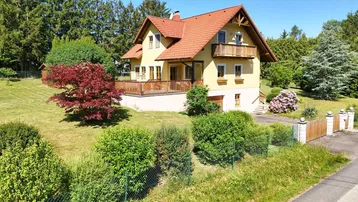 Gelbes Einfamilienhaus mit rotem Ziegeldach, umgeben von einem großen Garten und Bäumen unter blauem Himmel.