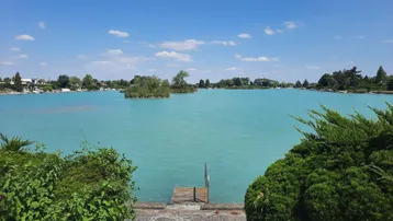 Weitläufiger Blick auf den türkisfarbenen See mit kleinem Holzsteg unter blauem Himmel.