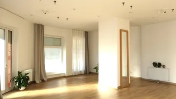 Heller, leerer Raum mit Holzboden, großen Fenstern und einem Wandspiegel, ideal für verschiedene Aktivitäten.