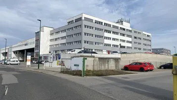Mehrstöckiges Gebäude mit moderner grau-weißer Fassade und umliegenden Parkplätzen unter bewölktem Himmel.
