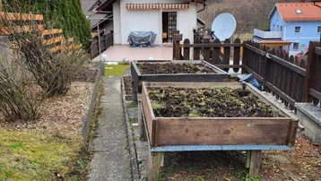 Gartenbereich mit Hochbeeten und einem gepflasterten Weg, umgeben von Hecken und Hügeln.
