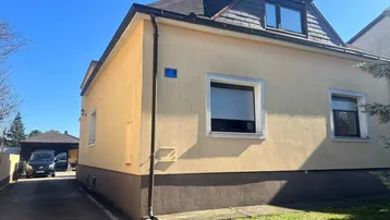 Seitenansicht des Hauses mit gelber Fassade, Rollläden und Einfahrt unter blauem Himmel.