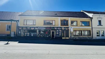 Gebäudeansicht mit gelber Fassade und einem Pizzeria-Geschäft im Erdgeschoss unter blauem Himmel.