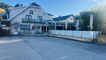 Das Gebäude präsentiert sich mit einer hellen Fassade, einem roten Ziegeldach und einem großen Parkplatz davor.